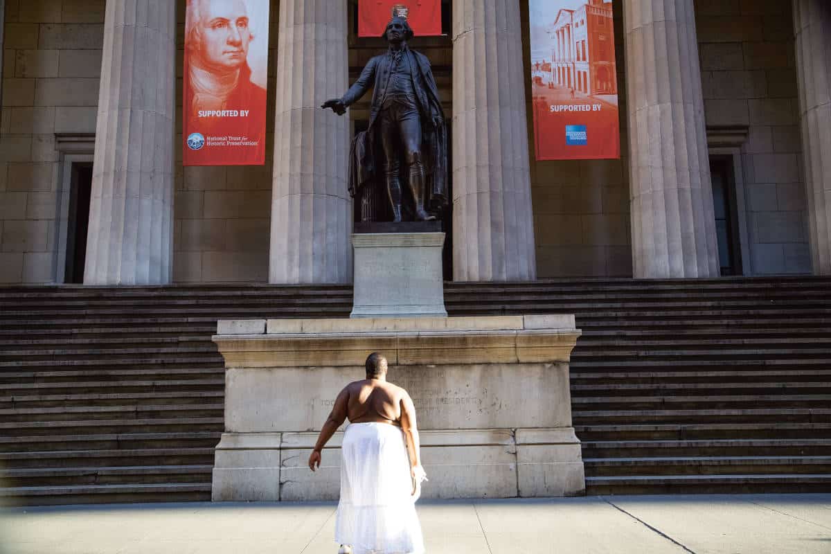 « ... a thirst for compleat freedom... had been her only motive for absconding, Oney Judge » (… sa soif de liberté totale… a été l’unique raison de sa fuite, Oney Judge), Federal Hall, NYC, 2016. Nona Faustine incarne Oney Judge (1773-1848), devant le Federal Hall de New York, en 2016. Réduite en esclavage par George Washington, Oney Judge est devenue servante de Martha Washington, l’épouse du premier président des États-Unis d’Amérique. Au début des années 1830, elle s’échappa de la maison présidentielle lorsqu’elle apprit que Martha Washington s’apprêtait à transférer son titre de propriété à sa petite-fille. Dans une interview de 1845, elle raconte son évasion : « Tandis que [les propriétaires] faisaient leurs bagages pour aller en Virginie, je faisais les miens pour partir, je ne savais pas où, car je savais que si je retournais en Virginie, je n’obtiendrais jamais ma liberté. J’avais des ami·es parmi les gens de couleur de Philadelphie, j’ai fait porter mes affaires à l’avance et j’ai quitté la maison des Washington pendant que la famille dînait. ». Photo : Nona Faustine. Crédit : Courtesy Nona Faustine and MACK.