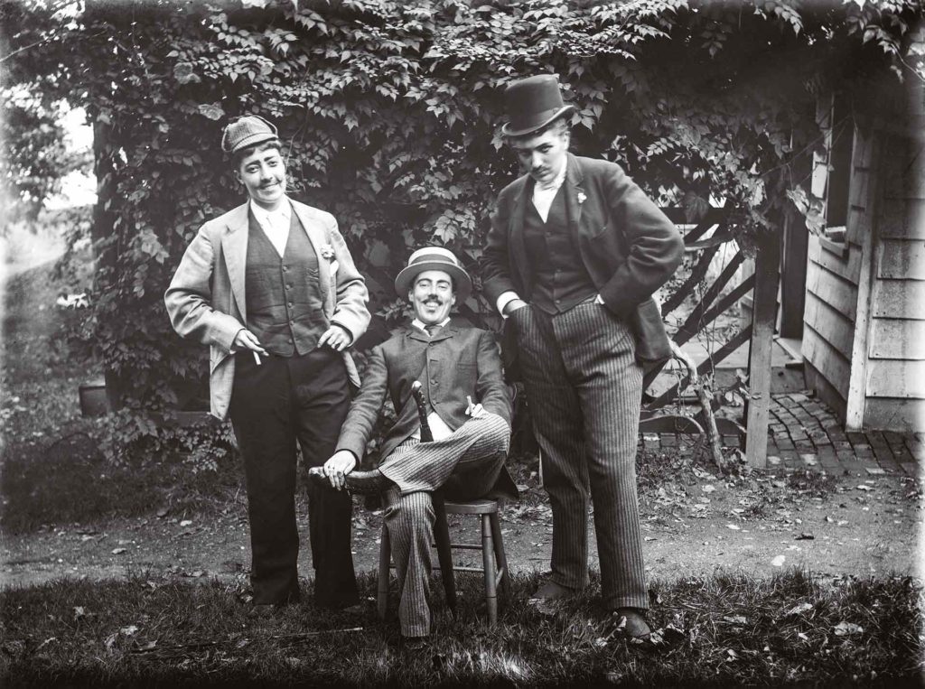 Julia Martin, Julia Bredt et moi-même en habit (1891). L’un des clichés les plus connus d’Alice Austen, qui pose au centre. Cette mise en scène des stéréotypes de la masculinité illustre son sens de l’humour subversif : elle tient, de manière suggestive, un parapluie entre les jambes.Crédit photo : Collection of Historic Richmond Town